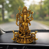 An ornate gold-toned Ganesha figurine displayed on a car dashboard with a blurred road and trees in the background.