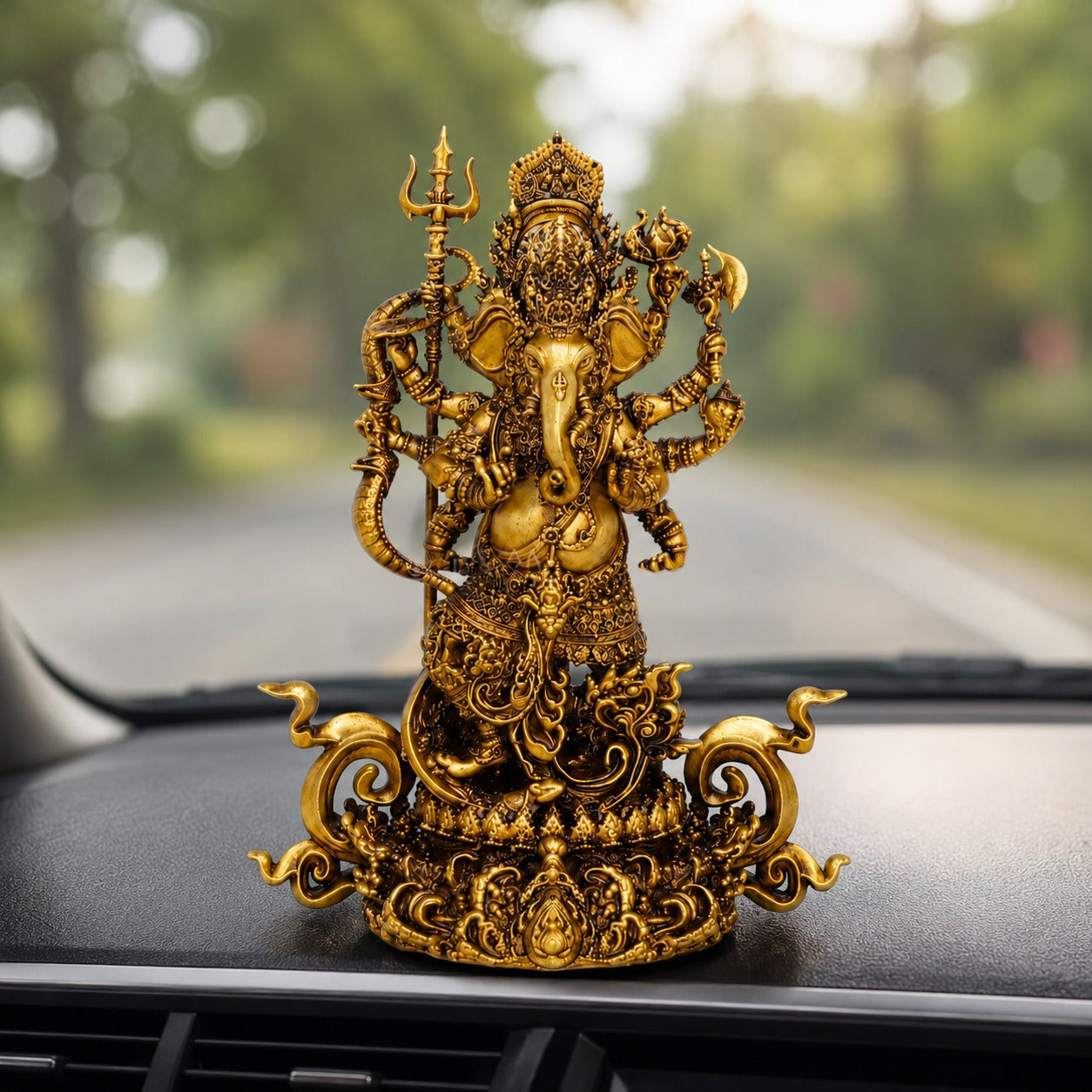 An ornate gold-toned Ganesha figurine displayed on a car dashboard with a blurred road and trees in the background.