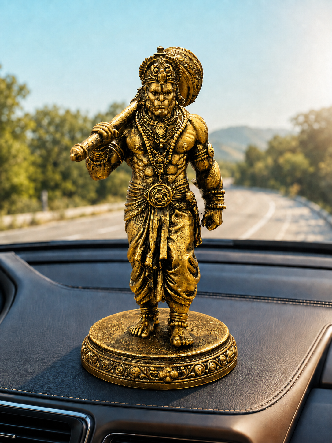 A golden brass Lord Hanuman statue standing on a round decorative base, placed on a car dashboard with a blurred road and trees in the background.